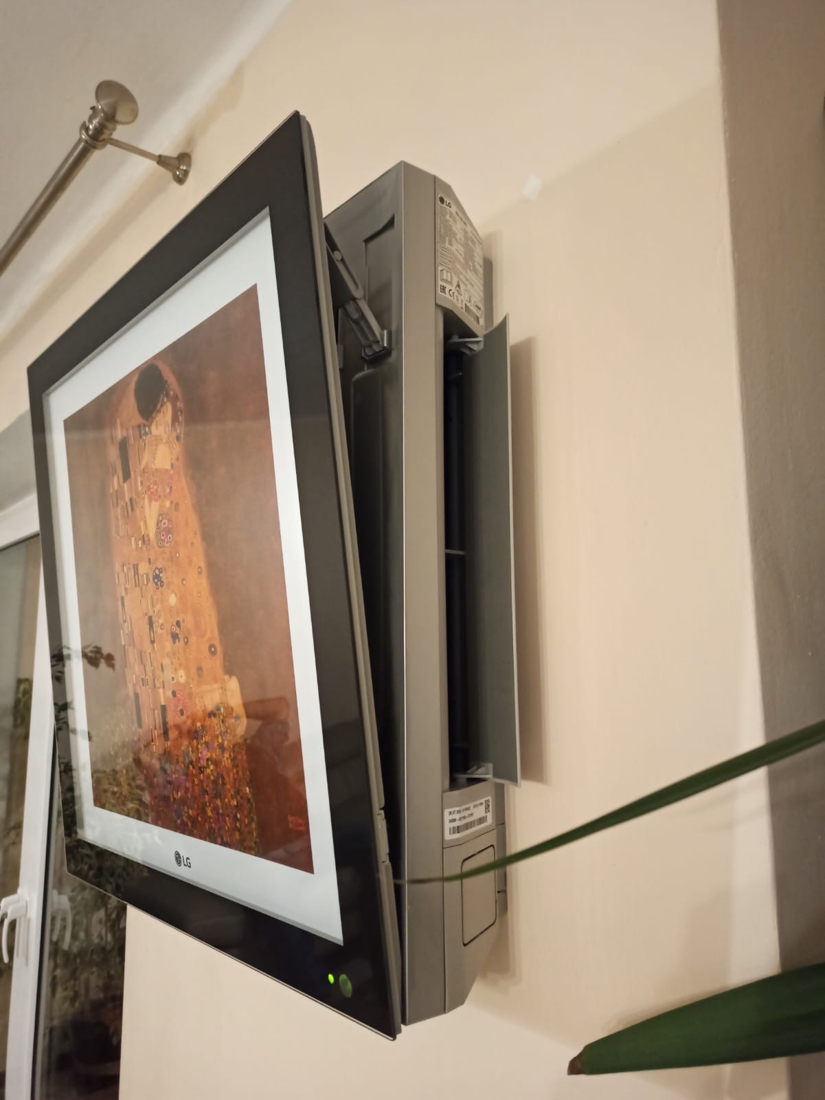 Klimatyzator ścienny LG Artcool Gallery z wyświetlanym obrazem 'Pocałunek' Gustava Klimta, zamontowany na beżowej ścianie, widoczne elementy montażowe i fragment rośliny doniczkowej.
