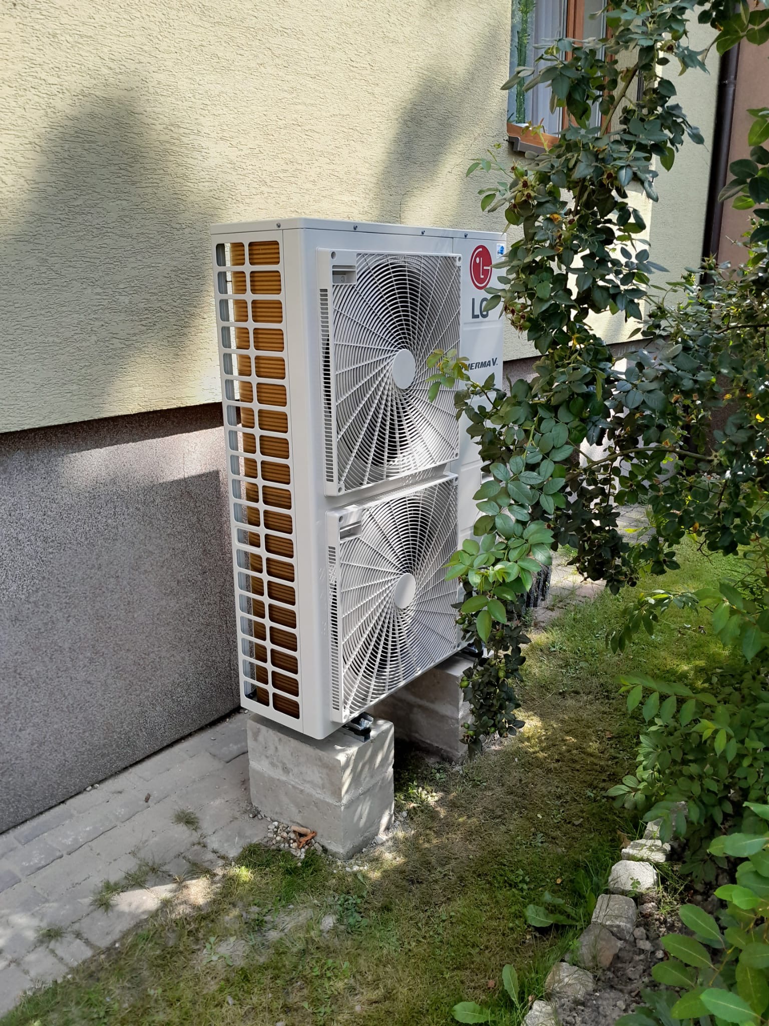 Zewnętrzna jednostka pompy ciepła LG Therma V, zamontowana na betonowych blokach przy ścianie domu z elewacją w kolorze beżowym, częściowo zasłonięta przez krzew z zielonymi liśćmi.