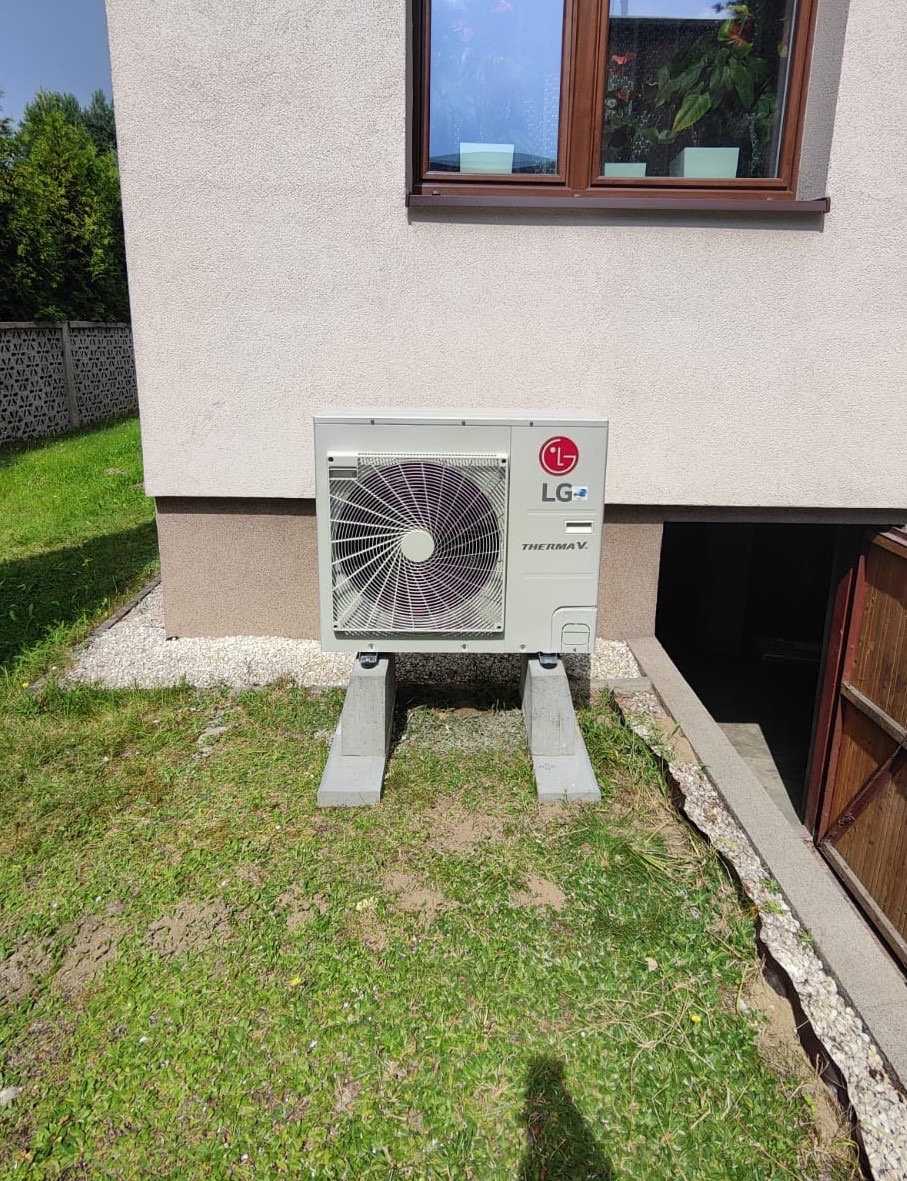 Zewnętrzna jednostka pompy ciepła LG Therma V zamontowana na betonowych wspornikach przy ścianie budynku z oknem i wejściem do piwnicy.