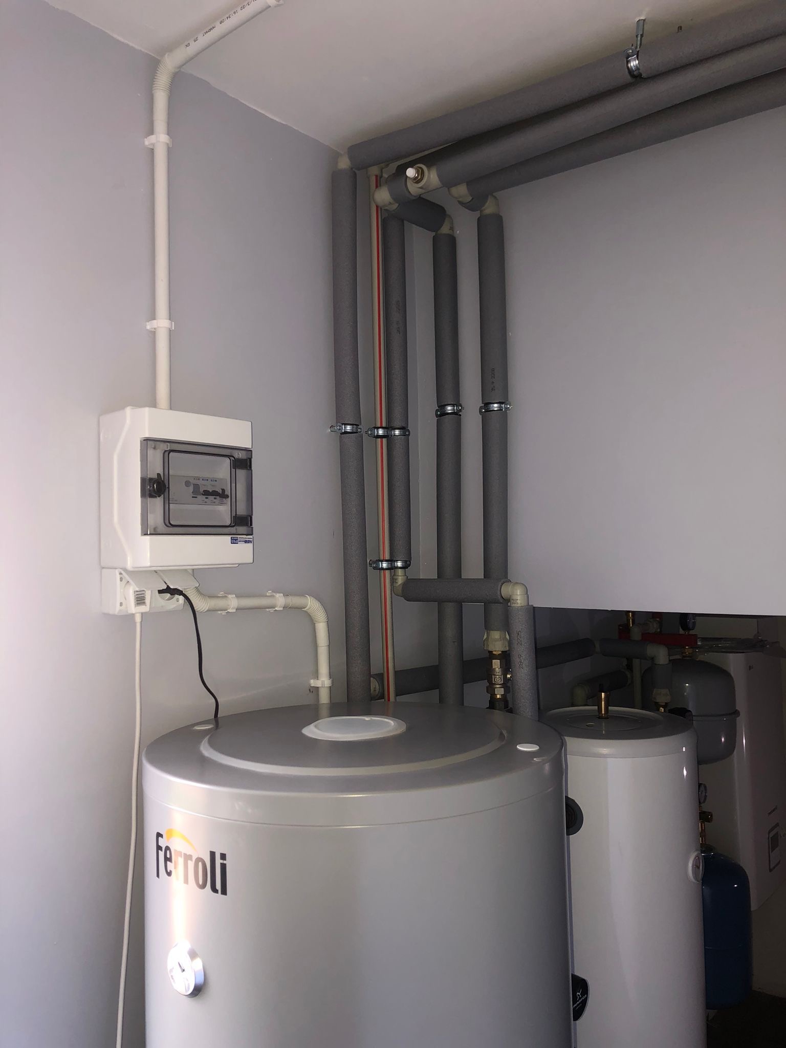 Szary zbiornik Ferroli z widocznym manometrem, instalacja rur w szarej izolacji, biała skrzynka elektryczna na szarej ścianie.