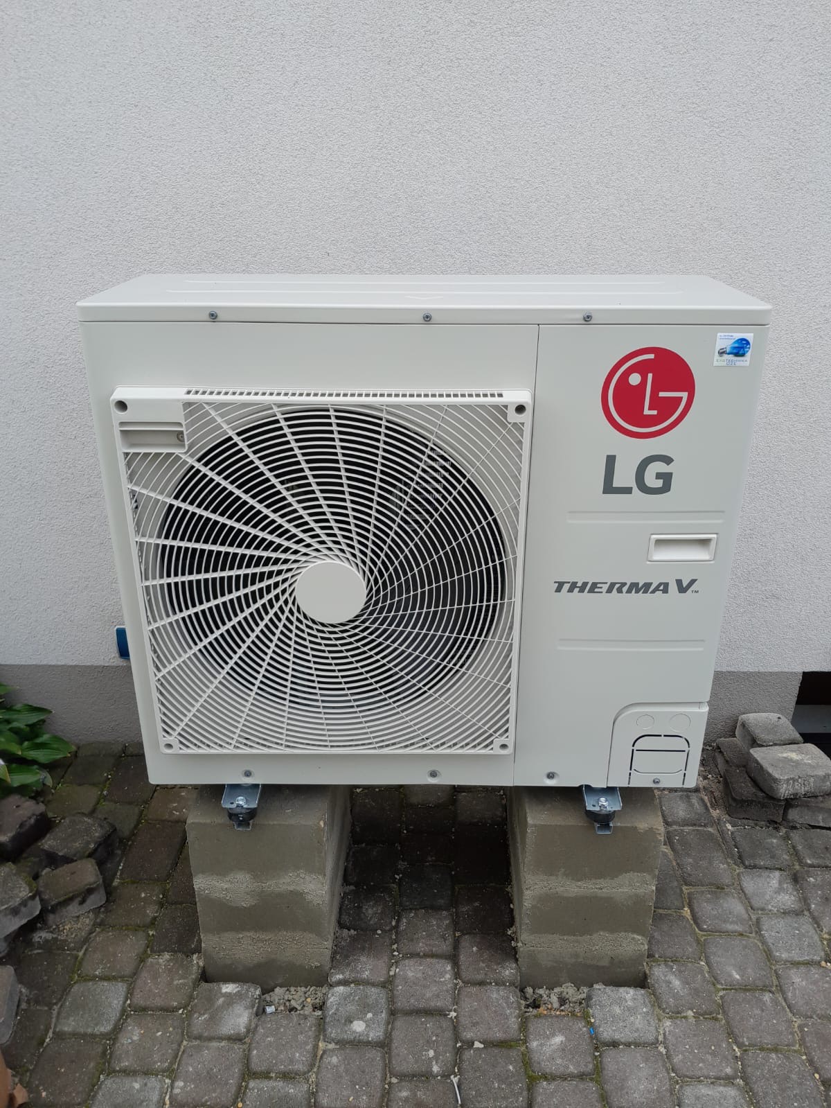 Zewnętrzna jednostka pompy ciepła LG Therma V, zamontowana na betonowych bloczkach na kostce brukowej, przy białej ścianie budynku.