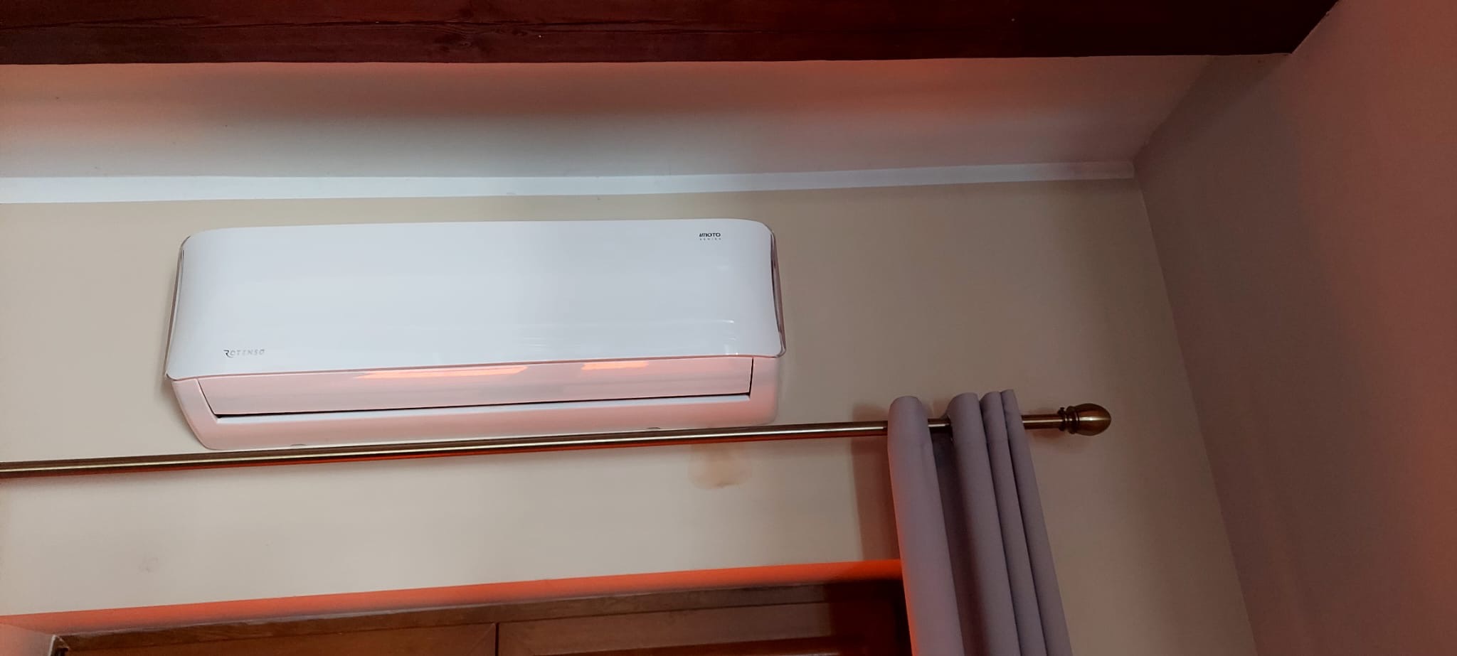 Biały klimatyzator marki Rotenso zamontowany na ścianie w pokoju, widoczny karnisz z zasłonami, oświetlenie tworzy ciepłą atmosferę.