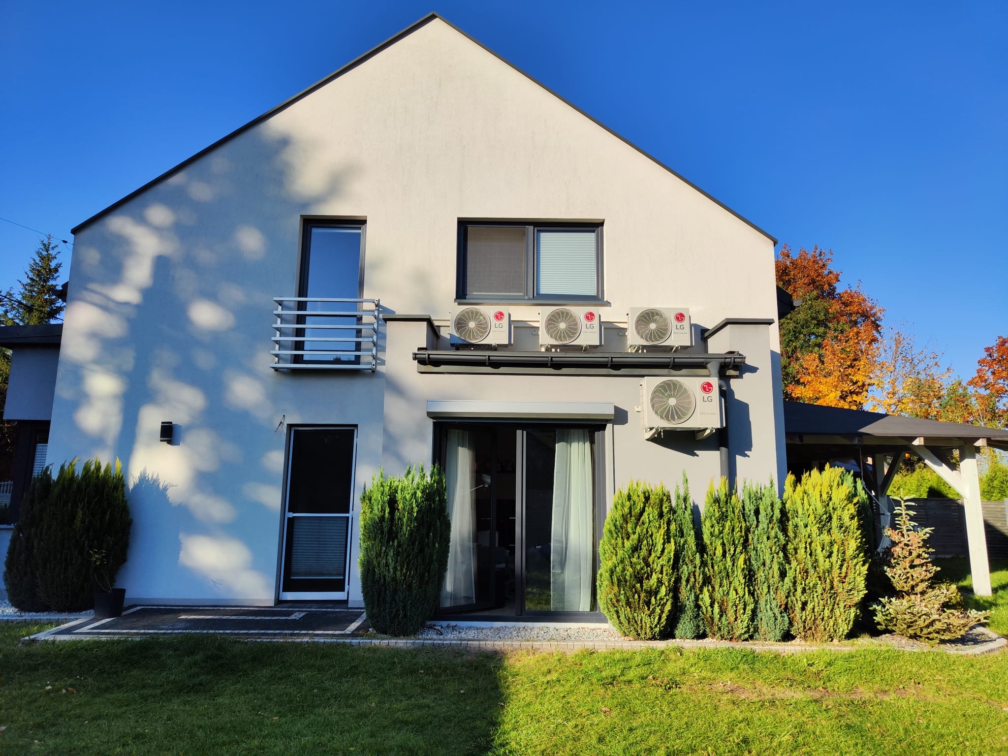 Elewacja domu jednorodzinnego z zamontowanymi czterema klimatyzatorami LG na specjalnej konstrukcji wsporczej, widoczne okna, balkon i zadbany ogród z tujami.