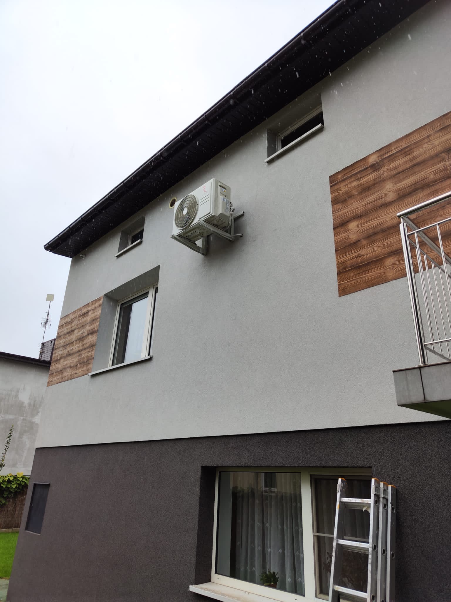 Zewnętrzna jednostka klimatyzacji zamontowana na szarej elewacji budynku, widoczna drabina oparta o ścianę, pochmurna pogoda.