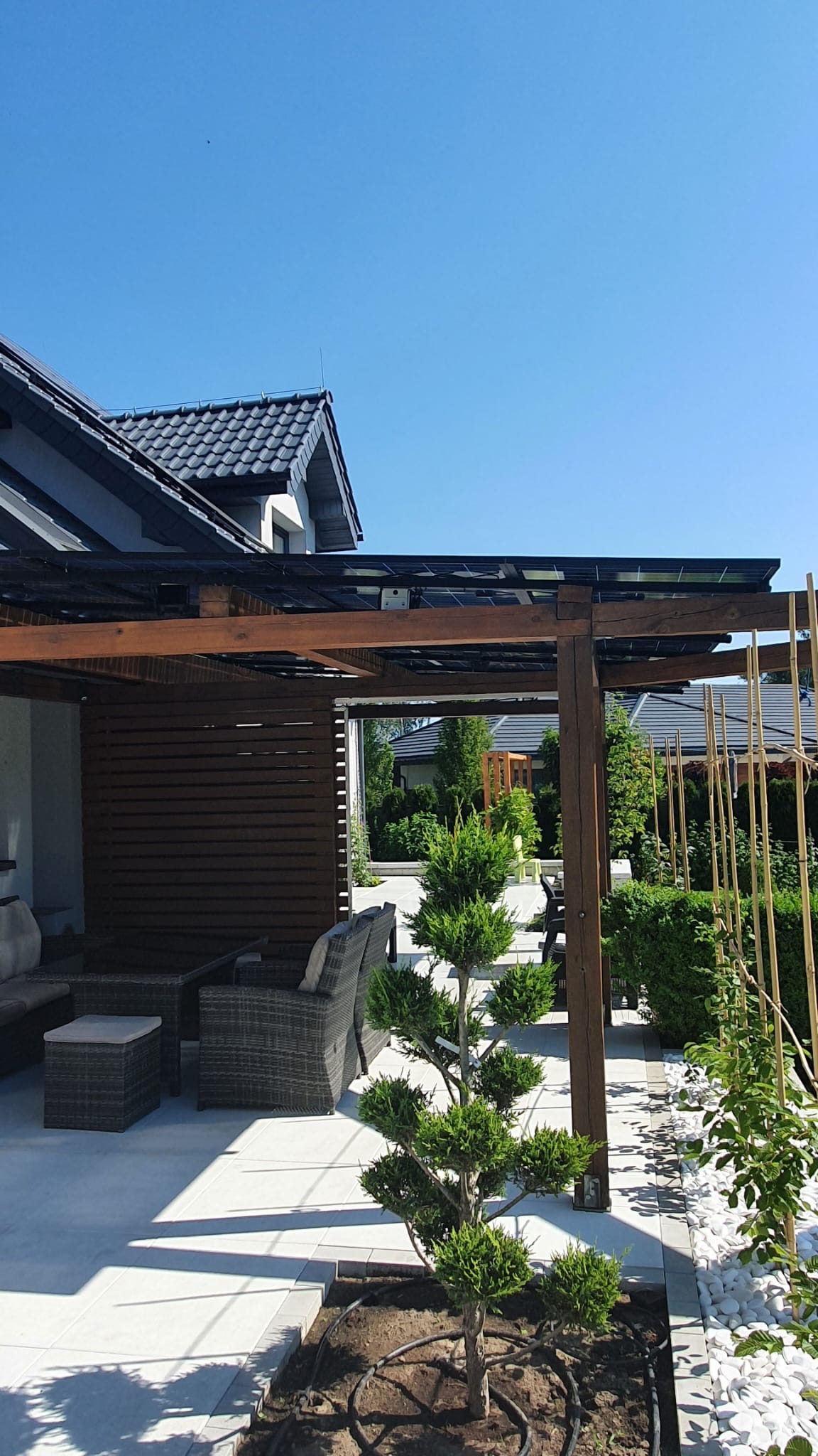 Nowoczesna pergola z panelami na dachu, otoczona zadbanym ogrodem i meblami ogrodowymi z rattanu.