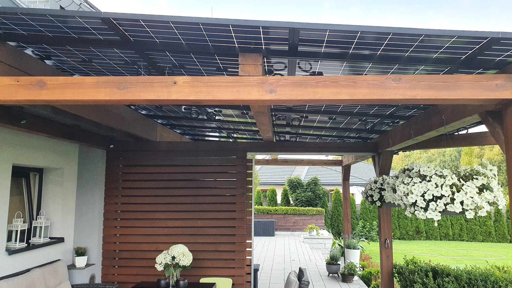 Zadaszenie tarasu z panelami słonecznymi, drewniana konstrukcja, widok na ogród z kwitnącymi kwiatami w wiszących donicach.