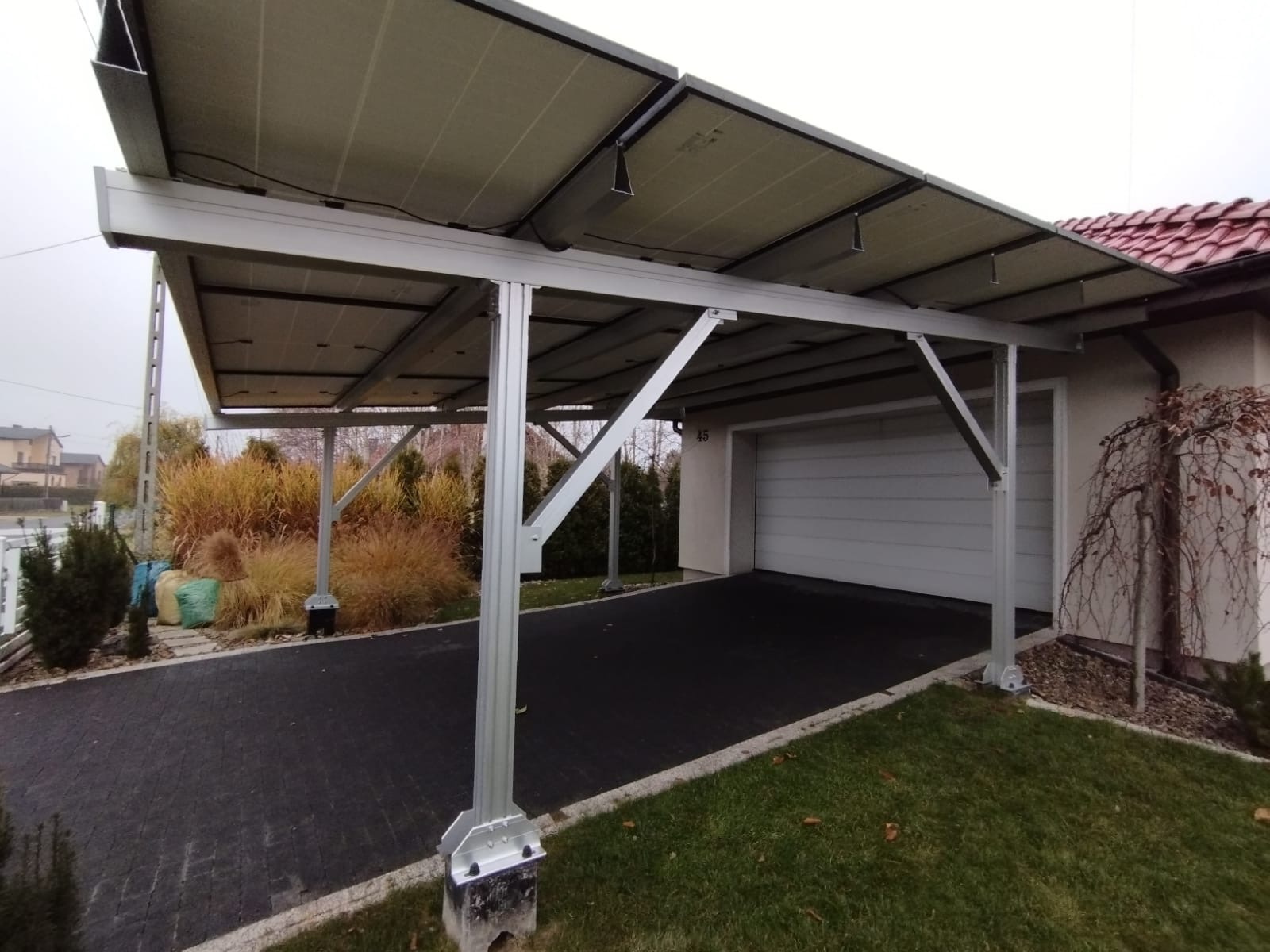 Zadaszenie wiaty garażowej z panelami fotowoltaicznymi, widok z zewnątrz na tle domu z garażem.