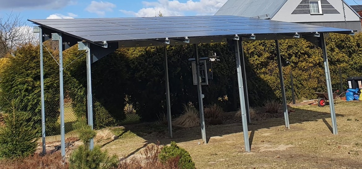 Konstrukcja wsporcza z panelami słonecznymi w ogrodzie, widoczne stalowe słupy nośne, trawnik, krzewy i dom w tle.