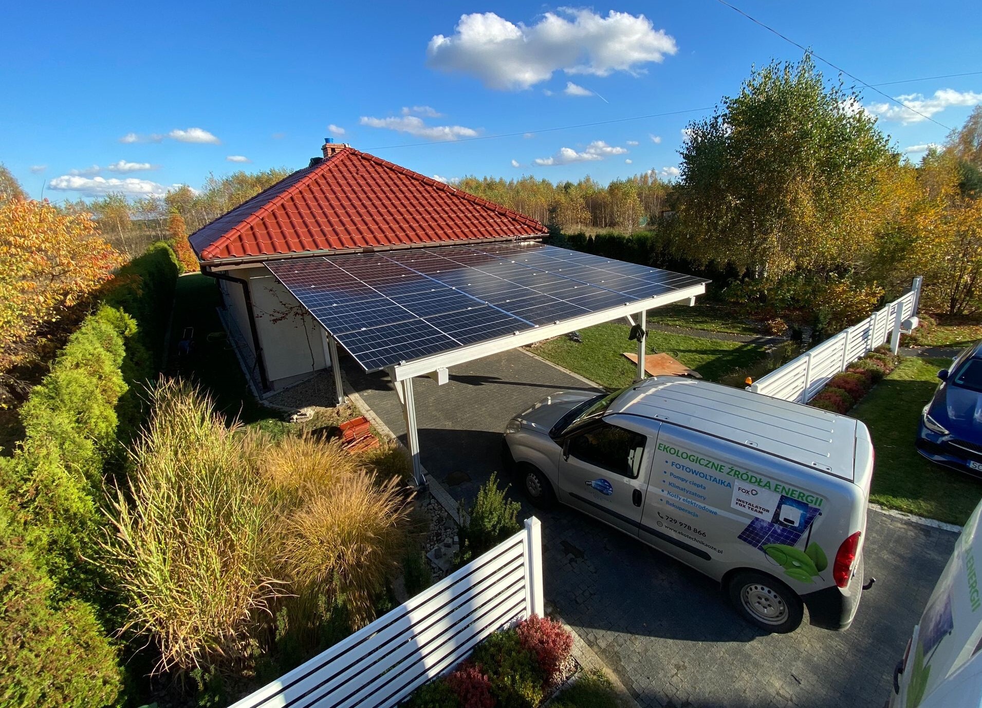 Dom z czerwonym dachem i panelami słonecznymi zamontowanymi nad wiatą garażową, widok z góry, obok zaparkowany srebrny samochód dostawczy z reklamą firmy instalującej panele fotowoltaiczne.