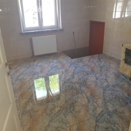 MJ Usługi remontowo budowlane - Wykończona podłoga z płytek imitujących marmur w jasnej kuchni z piecem kaflowym, widoczna instalacja grzewcza i okno z odbiciem drzew