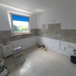 MJ Usługi remontowo budowlane - Wnętrze w trakcie prac w Zielonej Górze: częściowo wykończona ściana z szarymi płytkami imitującymi marmur, widoczne narzędzia i drabina, okno z niebieską roletą.