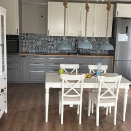 MJ Usługi remontowo budowlane - Wykończona kuchnia z szarymi szafkami dolnymi i białymi górnymi, wzorzystą ścianą nad blatem, drewnianym blatem, białym stołem z krzesłami oraz wiszącymi lampami z grubych lin.
