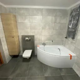 Nowoczesna łazienka po remoncie: narożna wanna z baterią, podwieszana toaleta ze spłuczką w zabudowie, wysoka szafka łazienkowa z jasnego drewna, ciemnoszare płytki podłogowe i jasnoszare płytki...