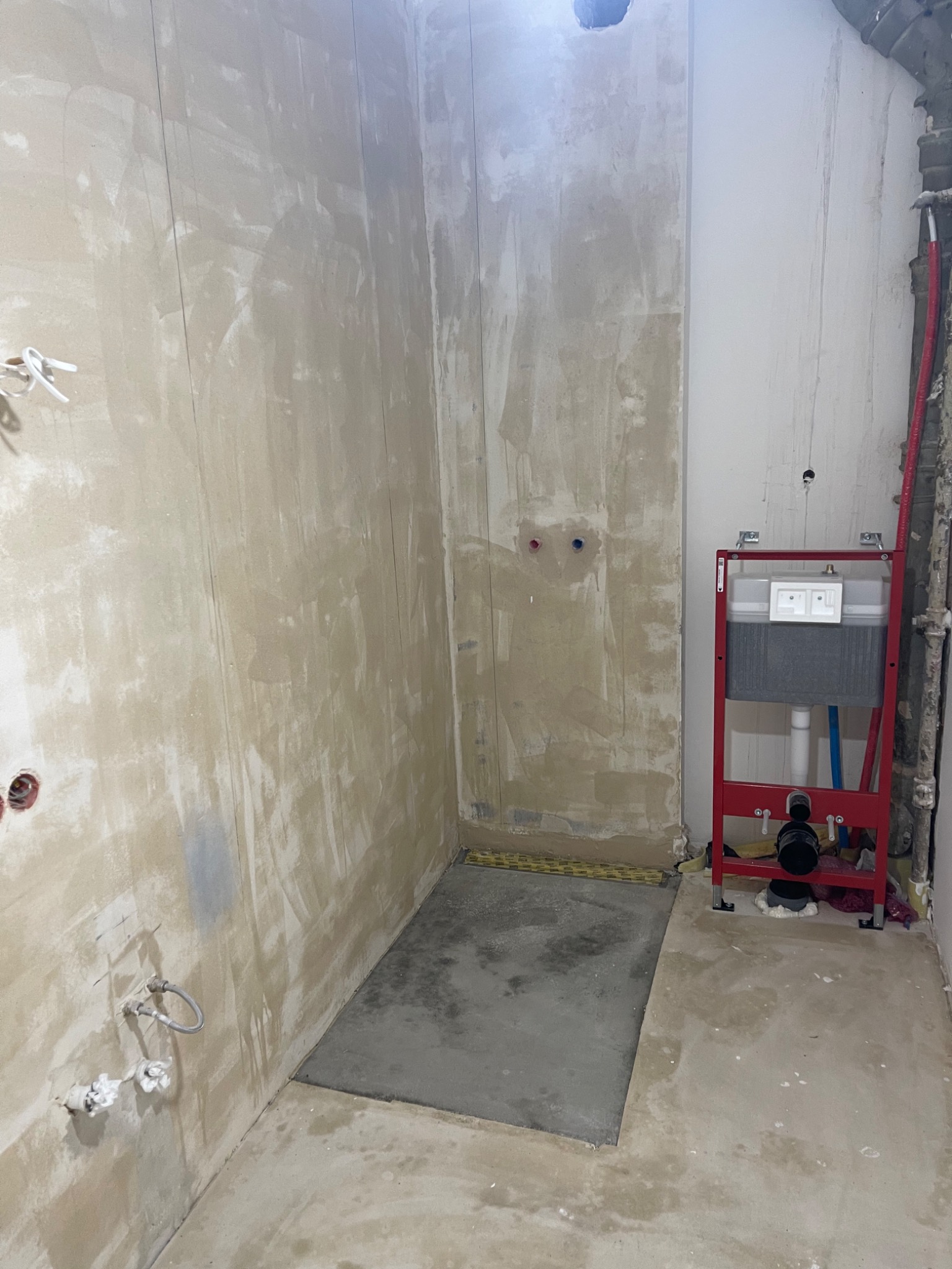 Nierówna ściana po gładziowaniu w trakcie remontu łazienki, widoczne instalacje hydrauliczne i stelaż WC podtynkowego w kolorze czerwonym.