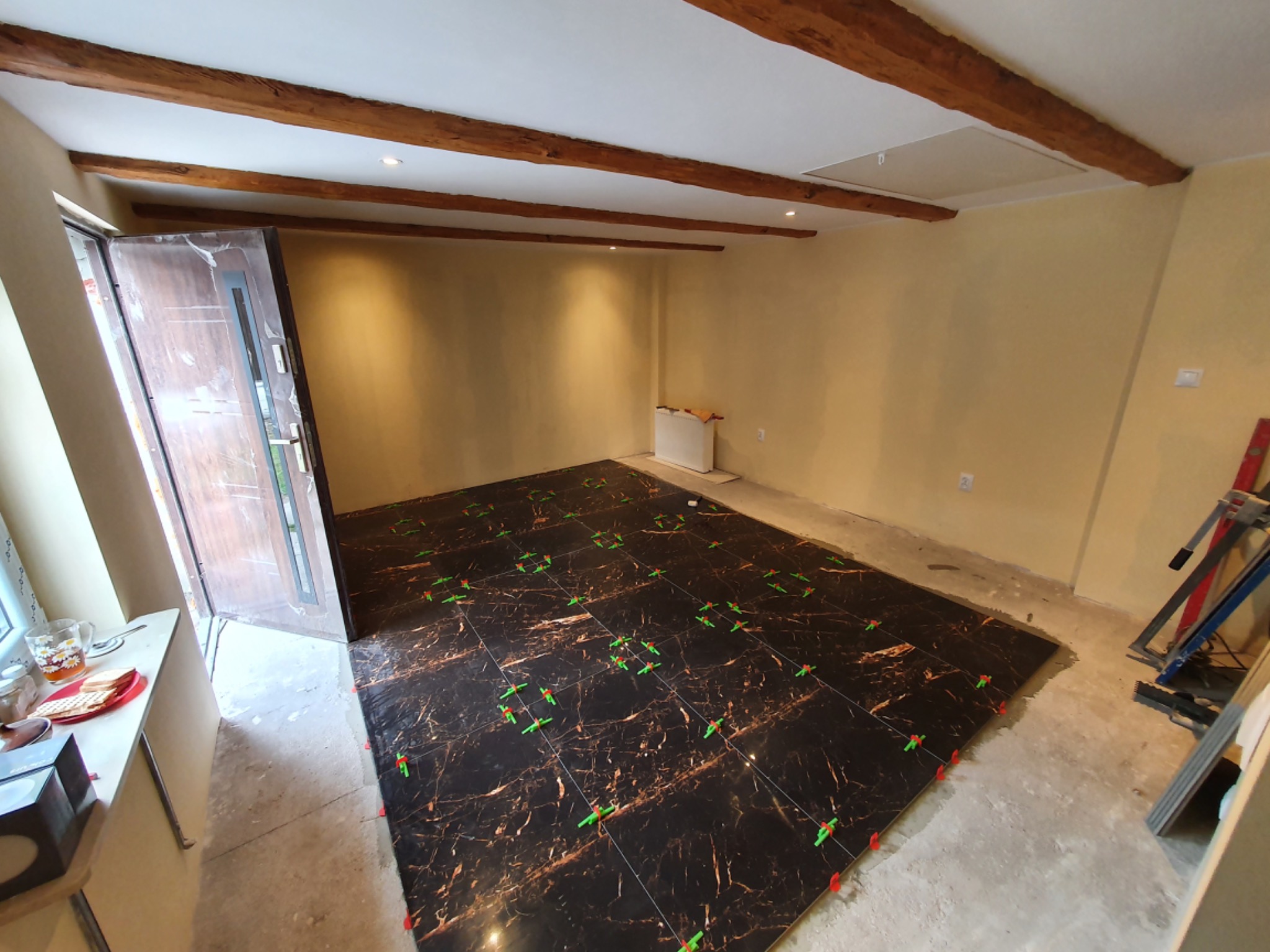 Częściowo ułożona, ciemna, marmurkowa podłoga z płytek, z zielonymi i czerwonymi klipsami poziomującymi, w pokoju z drewnianymi belkami stropowymi i otwartymi drzwiami.