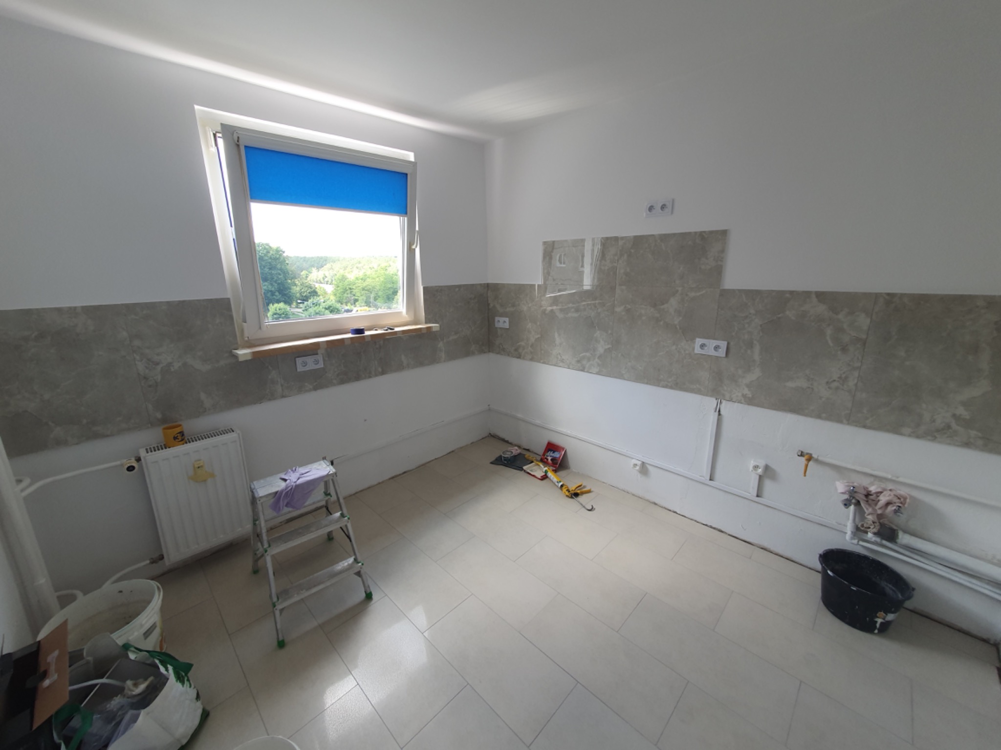 Wnętrze w trakcie prac w Zielonej Górze: częściowo wykończona ściana z szarymi płytkami imitującymi marmur, widoczne narzędzia i drabina, okno z niebieską roletą.