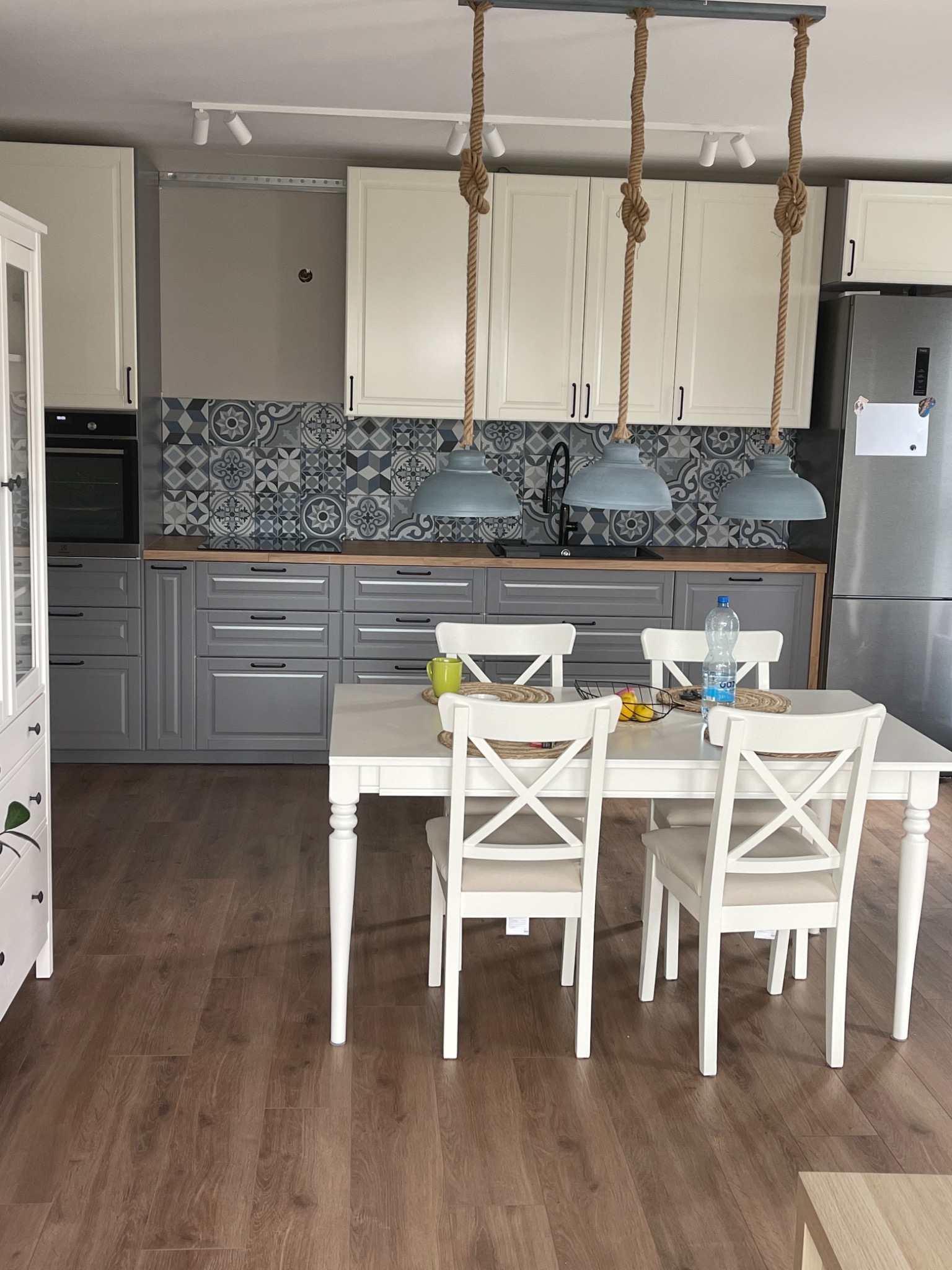 Wykończona kuchnia z szarymi szafkami dolnymi i białymi górnymi, wzorzystą ścianą nad blatem, drewnianym blatem, białym stołem z krzesłami oraz wiszącymi lampami z grubych lin.