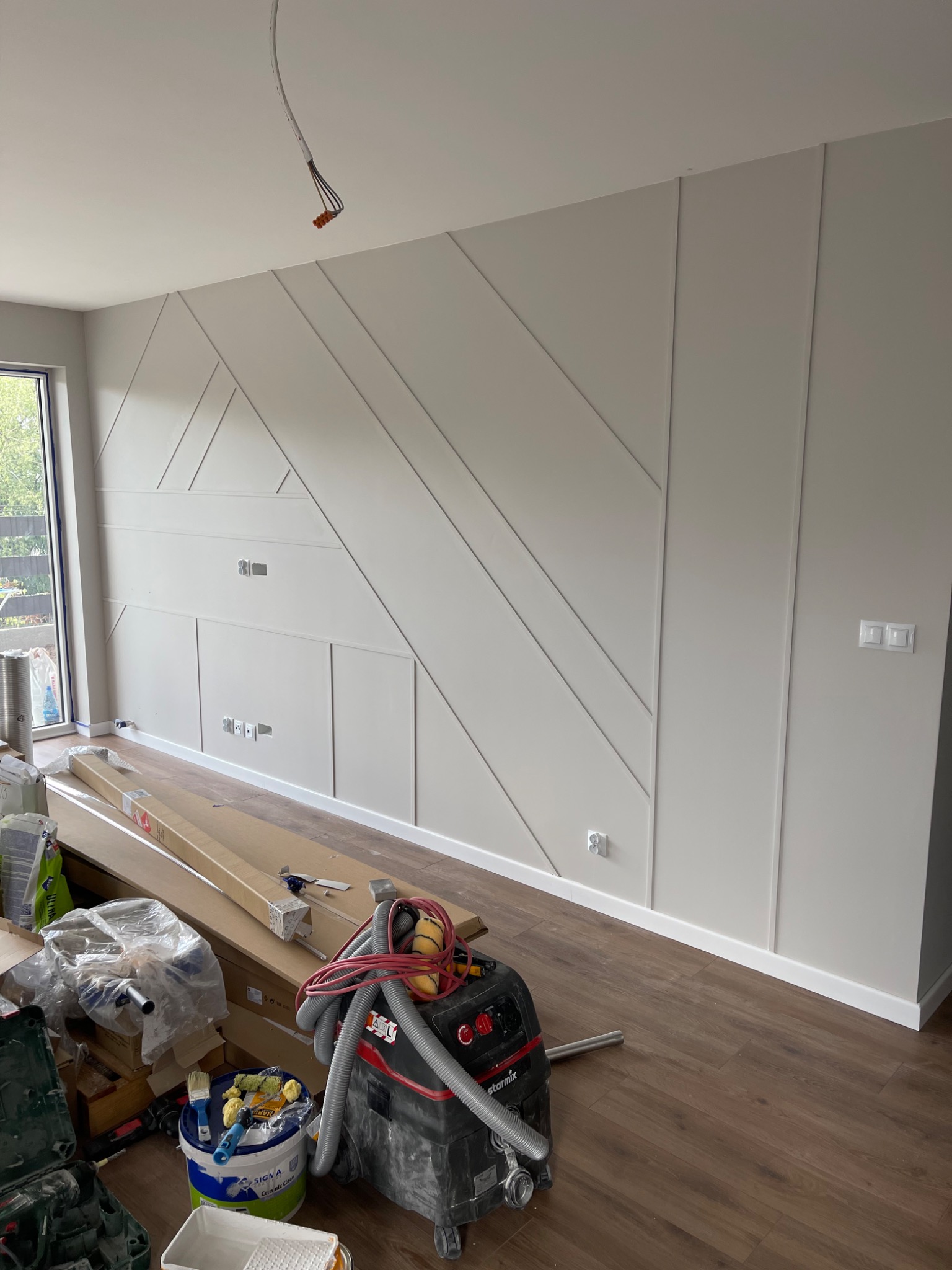 Ściana z dekoracyjnymi panelami ściennymi w trakcie montażu, widoczne narzędzia i materiały budowlane, drewnopodobna podłoga, gniazdka elektryczne w ścianie, okno z widokiem na zewnątrz.