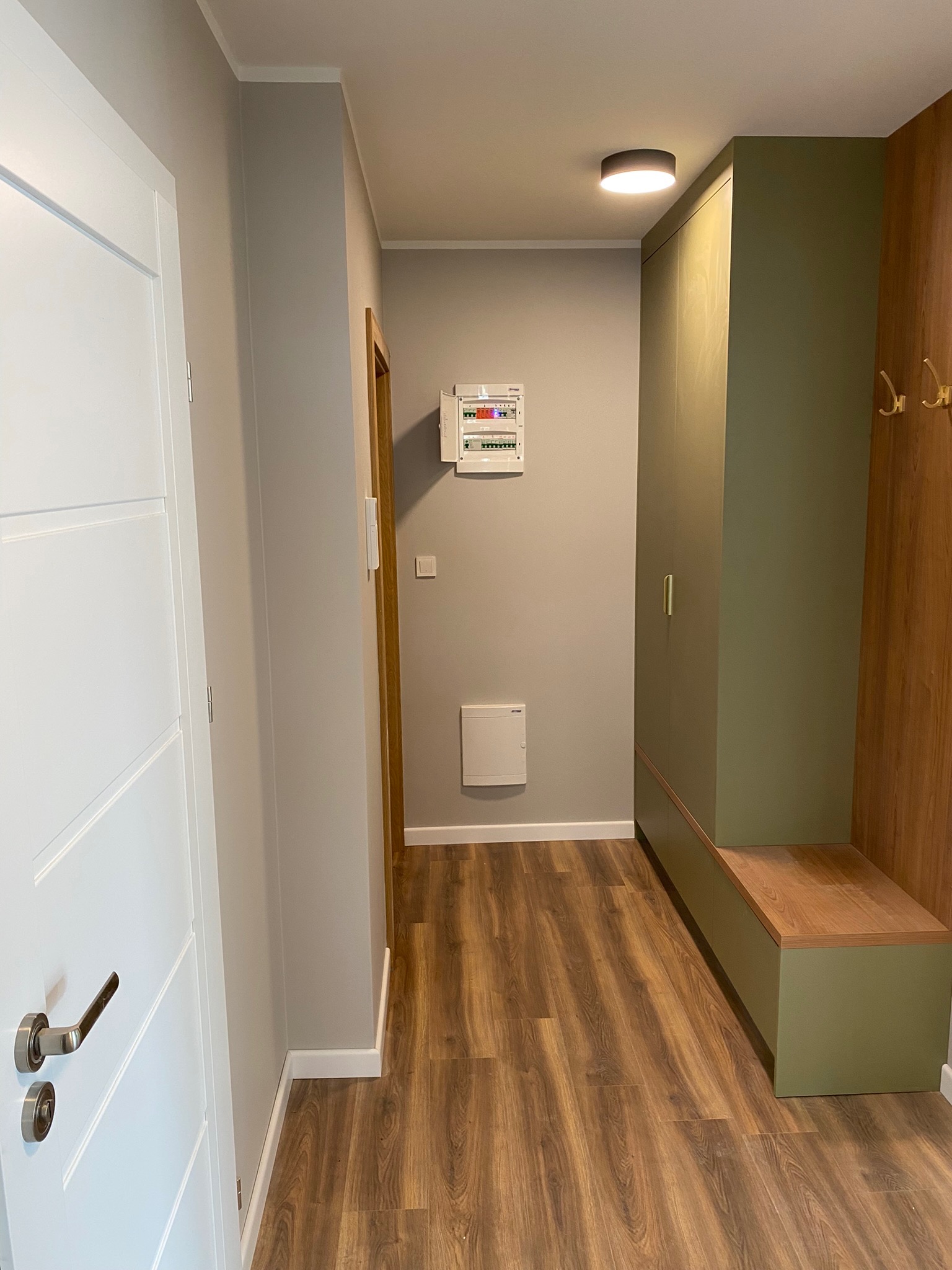 Nowoczesny przedpokój z drewnopodobną podłogą, szarą ścianą z białą skrzynką elektryczną, białą listwą przypodłogową, białymi drzwiami z srebrną klamką i zabudowaną szafą w kolorze oliwkowym...