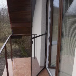 Otwarta moskitiera ramkowa w kolorze brązowym na balkonie z brązowymi płytkami, widoczna barierka i okno z żaluzjami.