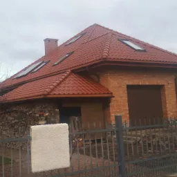 Dom jednorodzinny z nową, czerwoną dachówką ceramiczną z oknami dachowymi, widok zza szarego ogrodzenia.