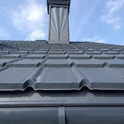 Nowoczesny, grafitowy dach z blachy trapezowej i oryginalnym kominem z motywem promieni, widok z dołu, błękitne niebo w tle.