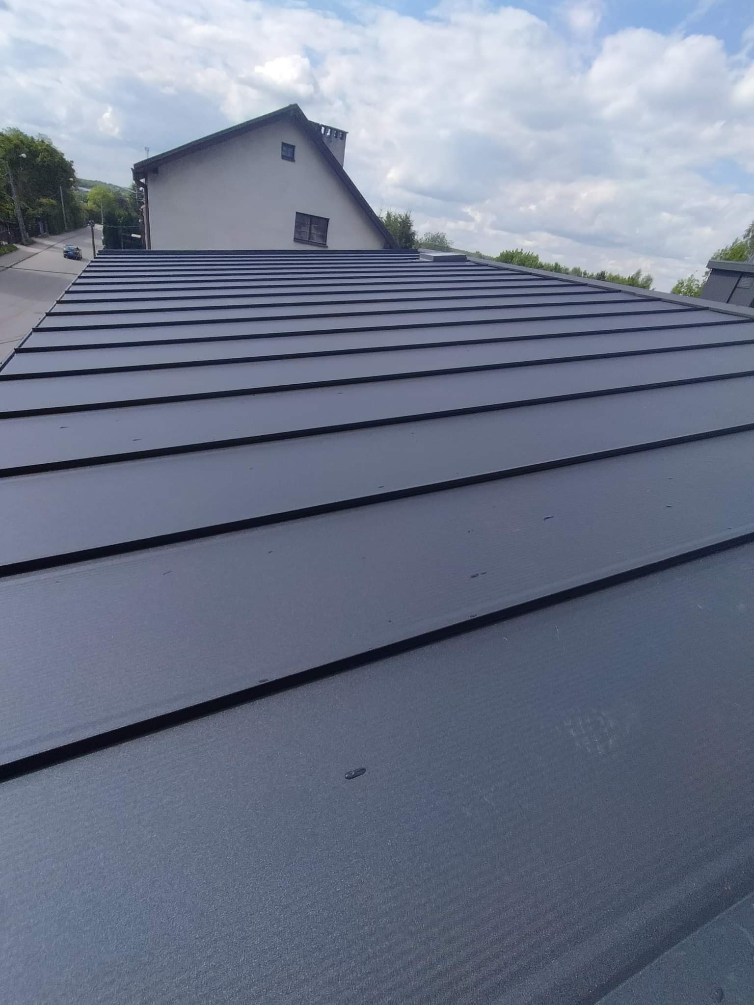 Nowy, grafitowy dach z blachy na domu jednorodzinnym, widok z góry, z widocznym fragmentem ulicy i niebem z chmurami.