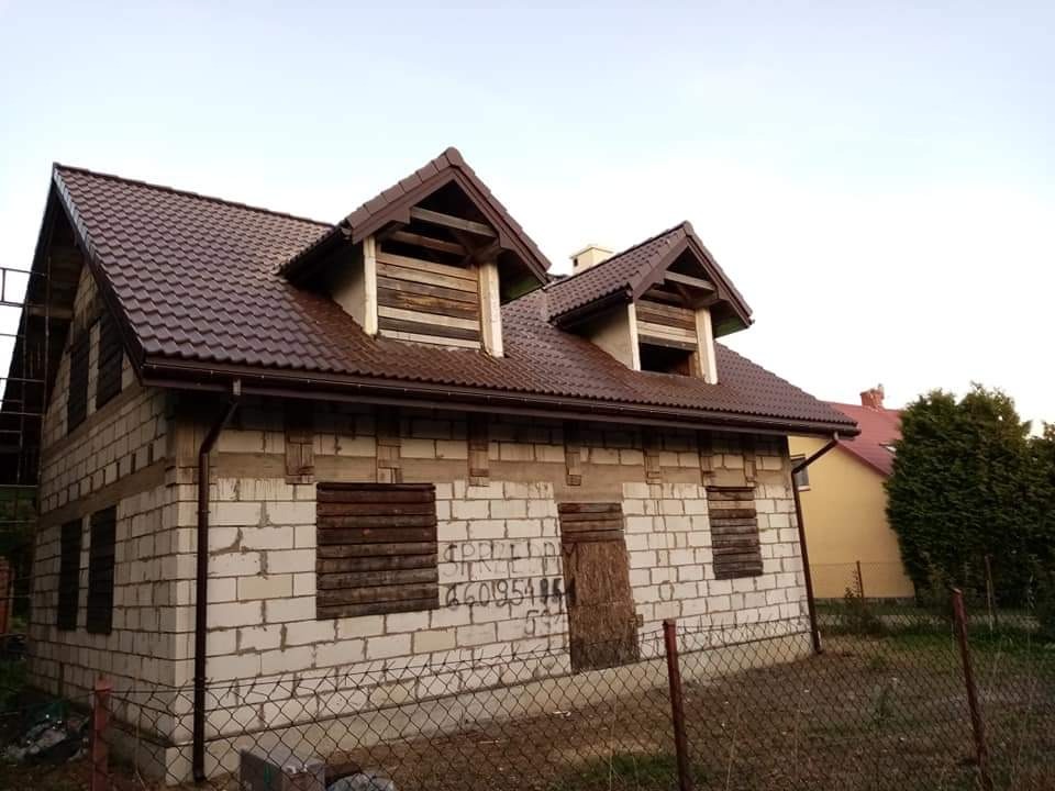 Surowy stan domu z białych bloczków z brązowym dachem z dachówki, zabezpieczone okna deskami, widoczne rusztowanie z boku budynku.