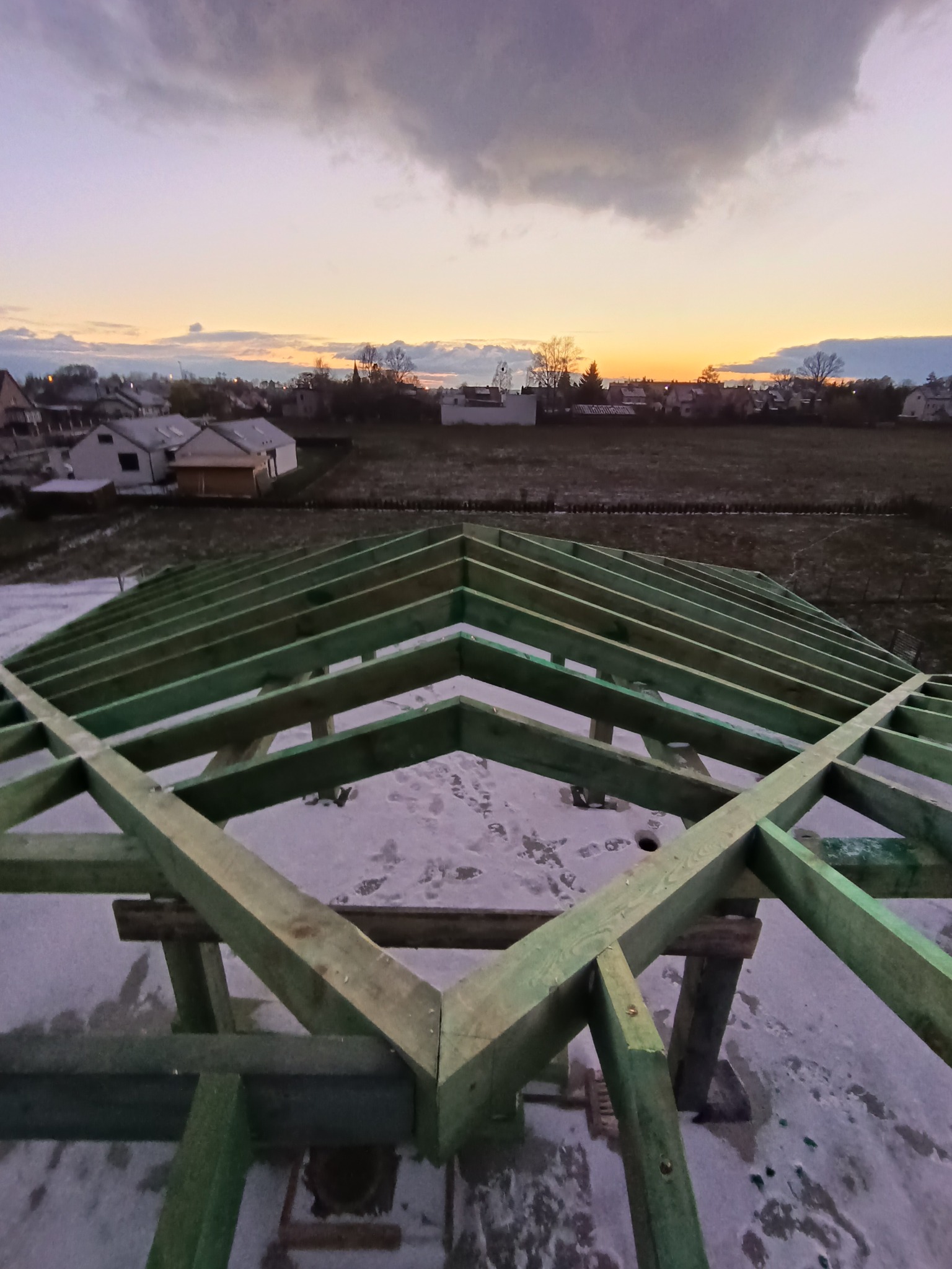 Surowa drewniana konstrukcja dachu w trakcie budowy, widok z góry, w tle zachodzące słońce i zabudowania.