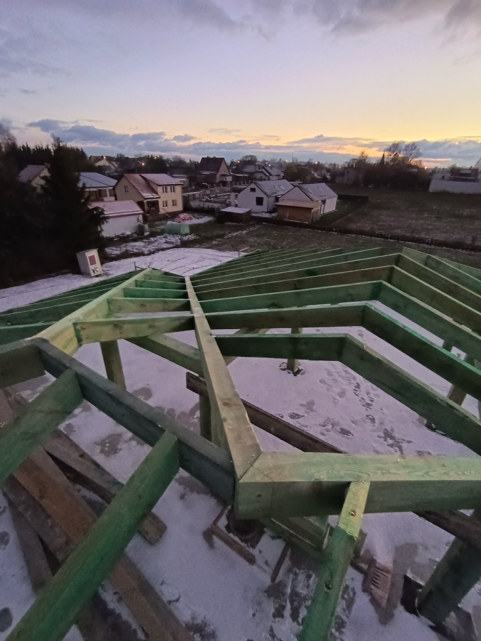 Surowa, zielona konstrukcja drewniana więźby dachowej na tle zaśnieżonego dachu i zabudowań mieszkalnych o zmierzchu.