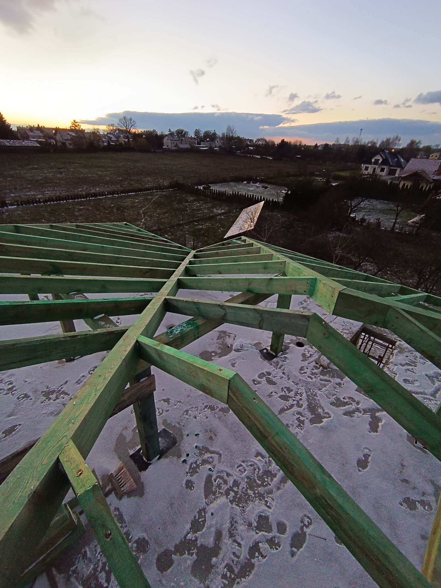 Konstrukcja dachu z zielonych belek drewnianych w trakcie montażu, widok z góry na tle zimowego krajobrazu o zmierzchu.