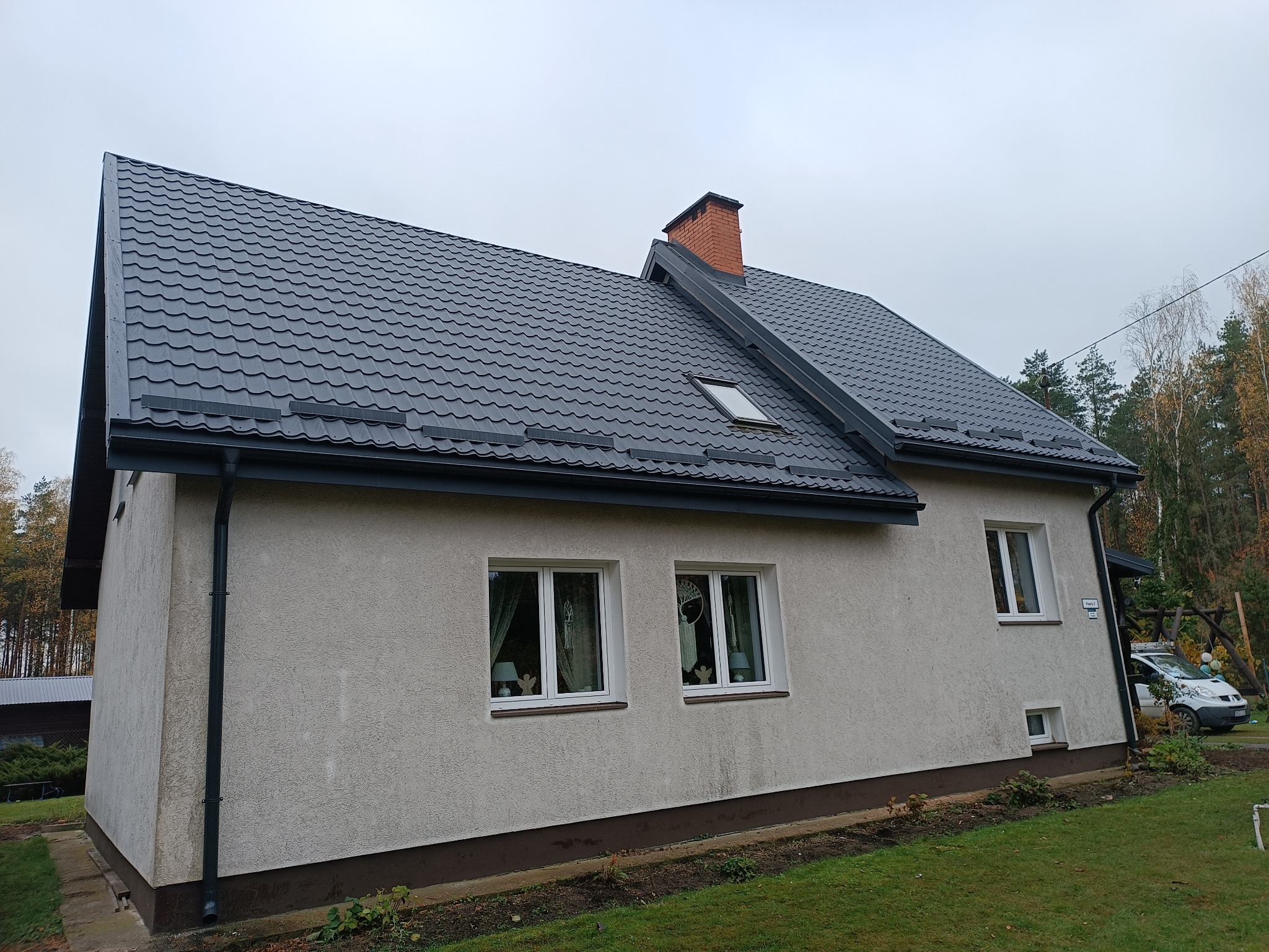Dom murowany z ciemnoszarą blachodachówką, ceglanym kominem i oknem dachowym, elewacja w odcieniach beżu, widoczne rynny i fragment ogrodu.