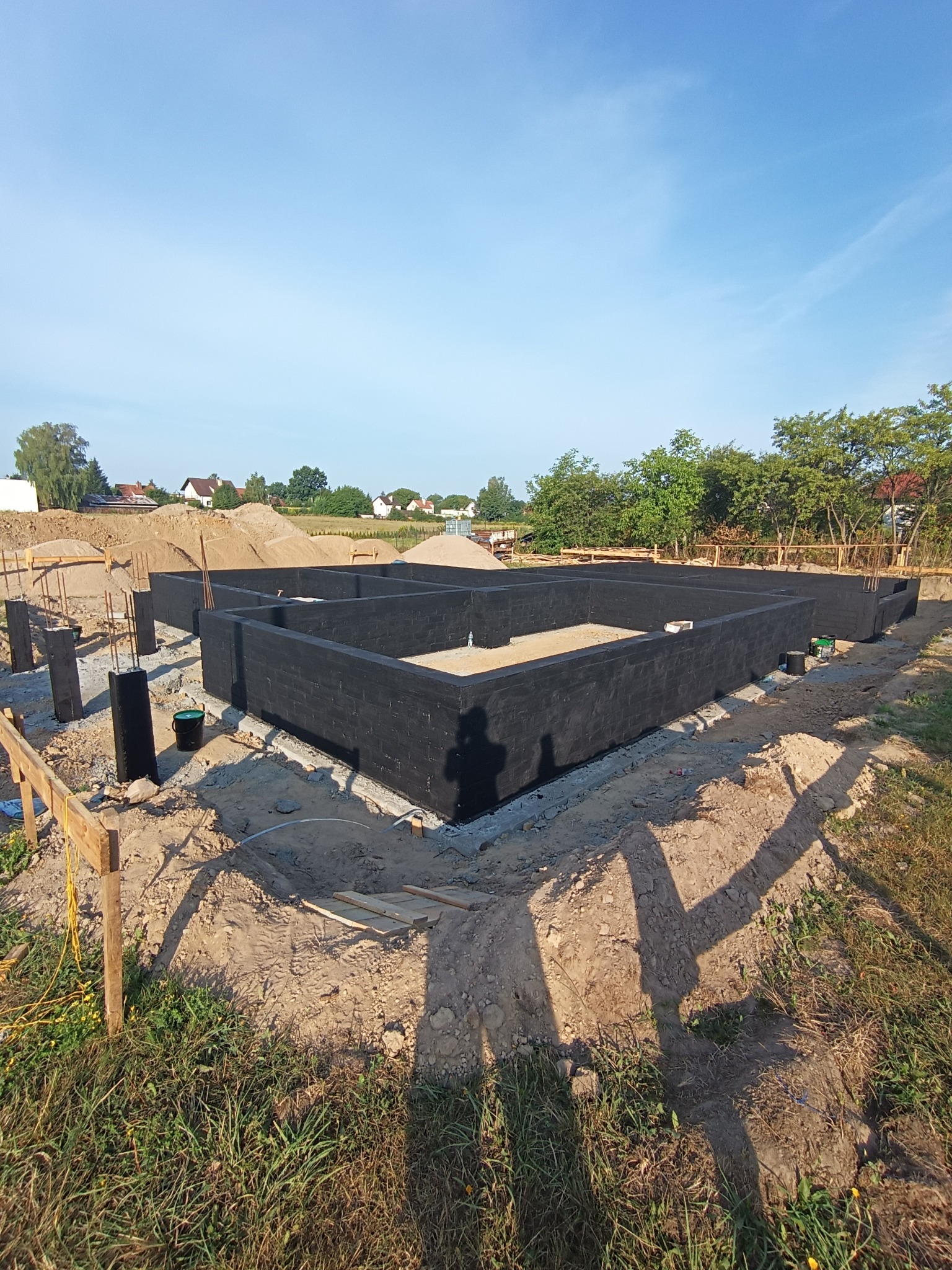 Fundamenty domu jednorodzinnego pomalowane na czarno, widoczne zbrojenia, teren budowy w słoneczny dzień.