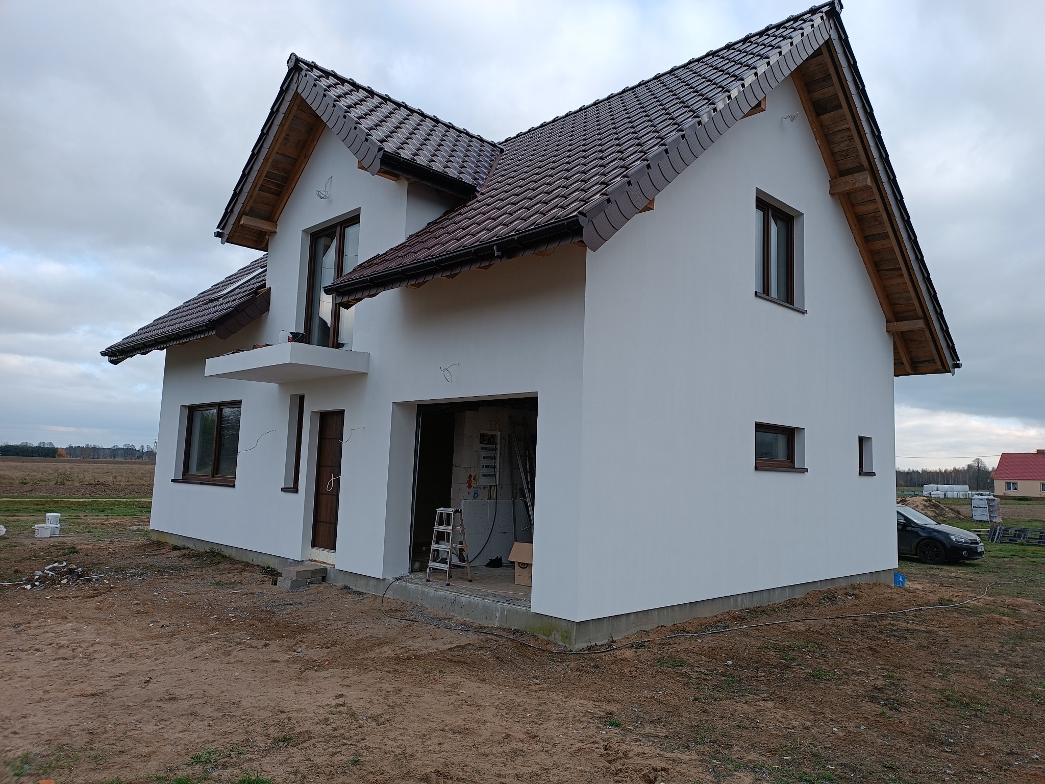 Dwukondygnacyjny dom murowany z brązowym dachem z dachówki ceramicznej, otynkowany na biało, z widoczną konstrukcją drewnianą dachu i niezadaszonym garażem w stanie surowym, z drabiną w środku,...