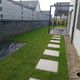 Nowoczesny ogród w Elblągu z trawnikiem, ścieżką z płyt betonowych i pergolą, widok na przygotowane podłoże pod nasadzenia przy ogrodzeniu.