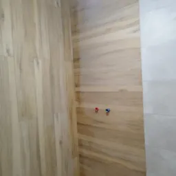 Ukończona ściana w łazience z płytek imitujących drewno, z widocznymi przyłączami wodnymi. Obok fragment ściany wyłożonej szarymi, prostokątnymi płytkami.