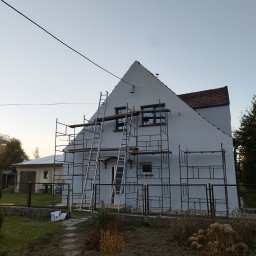 FIRMA BUDOWLANA ROMAN KOVALYK - Dom jednorodzinny w trakcie malowania elewacji, otoczony rusztowaniami i drabinkami. Widoczne puszki z farbą. Ujęcie z poziomu gruntu, w tle budynek gospodarczy.