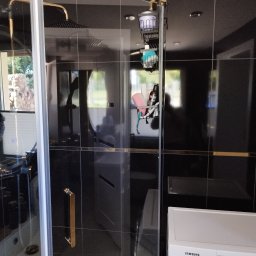 FIRMA BUDOWLANA ROMAN KOVALYK - Nowoczesna kabina prysznicowa z czarnymi płytkami i złotymi akcentami. Widoczna armatura w kolorze złota i biała pralka. Minimalistyczny design łazienki.