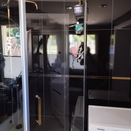 FIRMA BUDOWLANA ROMAN KOVALYK - Kabina prysznicowa z czarnymi płytkami i złotymi akcentami. Widoczna armatura, półki z kosmetykami i pralka. Nowoczesny design łazienki.