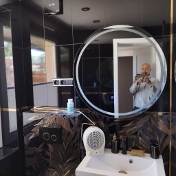 FIRMA BUDOWLANA ROMAN KOVALYK - Wykończona łazienka w czerni i złocie: czarne płytki, umywalka z szafką, okrągłe lustro z podświetleniem, dekoracyjne wzory na ścianach. Widoczne gniazdka i wentylator.