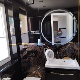 FIRMA BUDOWLANA ROMAN KOVALYK - Elegancka, czarno-złota łazienka z okrągłym lustrem podświetlanym LED, umywalką w szafce i toaletą. Widoczne czarne płytki z motywem liści i grzejnik elektryczny.