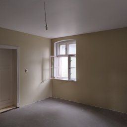 FIRMA BUDOWLANA ROMAN KOVALYK - Pomalowane na beżowo wnętrze pokoju z otwartym oknem i szarą podłogą. Widoczne kable zwisające z sufitu. Świeżo odnowione ściany i podłoga.