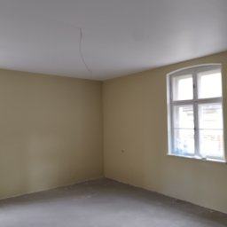 FIRMA BUDOWLANA ROMAN KOVALYK - Pomalowane na beżowo wnętrze pokoju z oknem. Widoczne ściany, sufit z kablem i szara podłoga. Świeżo po malowaniu, gotowe do dalszych prac wykończeniowych.