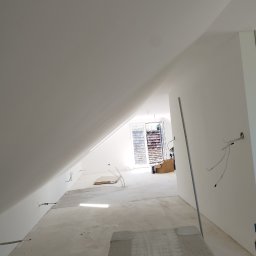 FIRMA BUDOWLANA ROMAN KOVALYK - Jasne poddasze po gładziowaniu ścian i sufitu, widoczne kable elektryczne, okno balkonowe w tle. Minimalistyczne wykończenie wnętrza w bieli.