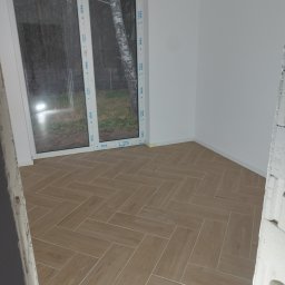 FIRMA BUDOWLANA ROMAN KOVALYK - Ukończona podłoga z płytek ułożonych w jodełkę w jasnym pokoju z dużym oknem. Widoczne białe ściany i fragment surowego muru. Płytki imitują drewno.