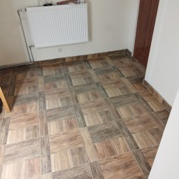 FIRMA BUDOWLANA ROMAN KOVALYK - Ukończona podłoga z płytek imitujących drewno, ułożonych w kwadraty, z grzejnikiem na ścianie i brązowymi listwami przypodłogowymi. Widok na fragment pomieszczenia.