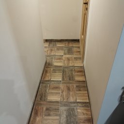 FIRMA BUDOWLANA ROMAN KOVALYK - Ułożona, brązowo-beżowa podłoga z płytek w wąskim korytarzu. Widoczna drewniana ościeżnica drzwi i fragment ściany w odcieniach beżu i szarości. Nowoczesne wykończenie.