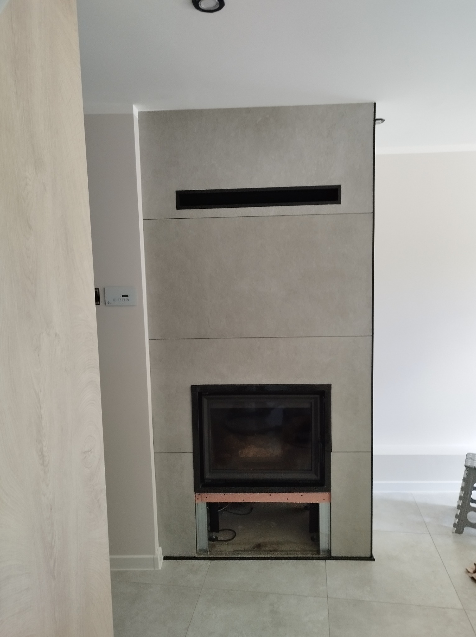 Obudowa kominka z szarych płyt, z czarną ramą wkładu i poziomymi szczelinami wentylacyjnymi. Widoczny fragment podłogi z szarych płytek i fragment ściany.