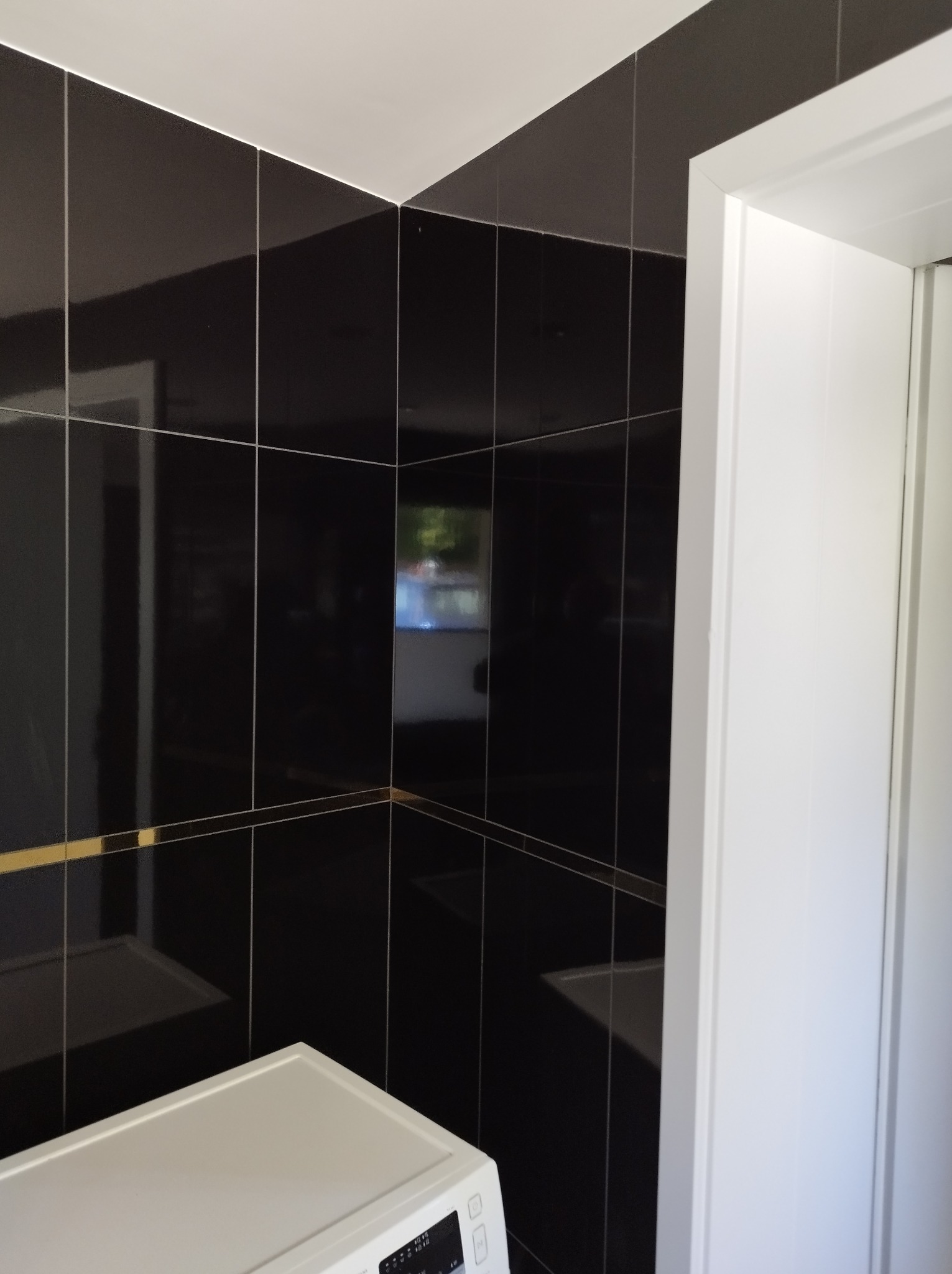 Czarne, błyszczące płytki z cienką, złotą fugą na ścianie łazienki, widoczne zza białej ramy drzwi i fragmentu pralki. Minimalistyczny design.