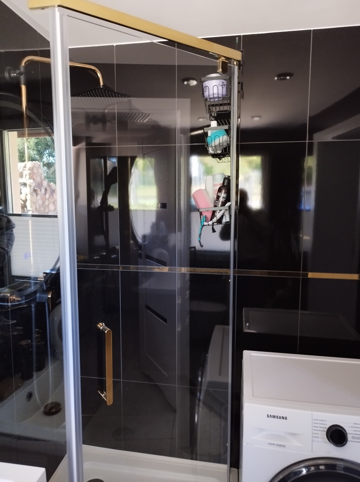 Kabina prysznicowa z czarnymi płytkami i złotymi akcentami. Widoczna armatura, półki z kosmetykami i pralka. Nowoczesny design łazienki.